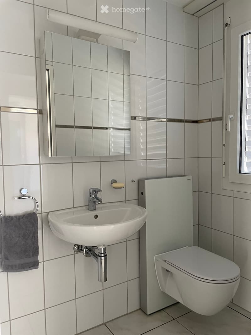 Modern bathroom with white tiles, a sink with a chrome faucet, a toilet, a large mirror, and a towel on a ring.