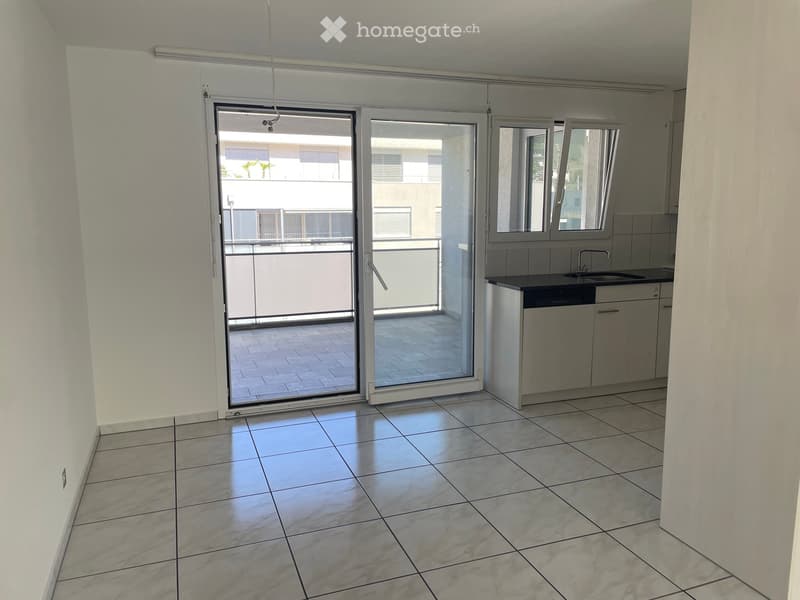 Empty room with tiled floor, white walls, sliding glass doors leading to a balcony, and a small kitchen area with sink and cabinets.