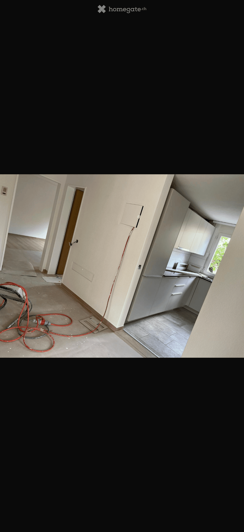 Empty room with unfinished walls and floor, visible electrical wiring, and a small kitchen area with cabinets and sink.
