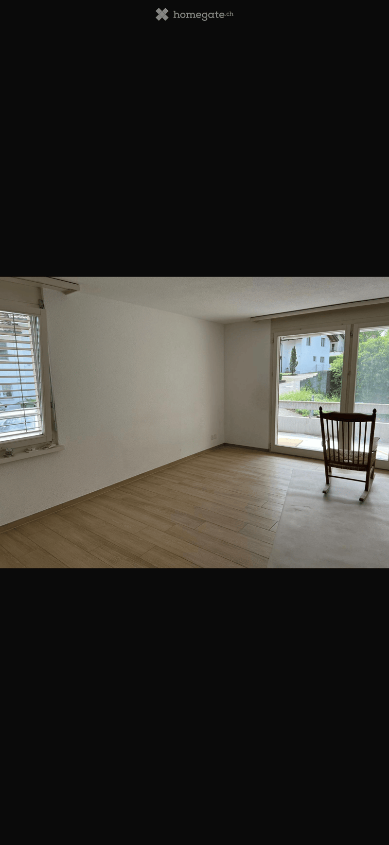 Empty room with wooden flooring, white walls, large window with blinds, and a single wooden chair near the sliding glass door leading to a balcony.