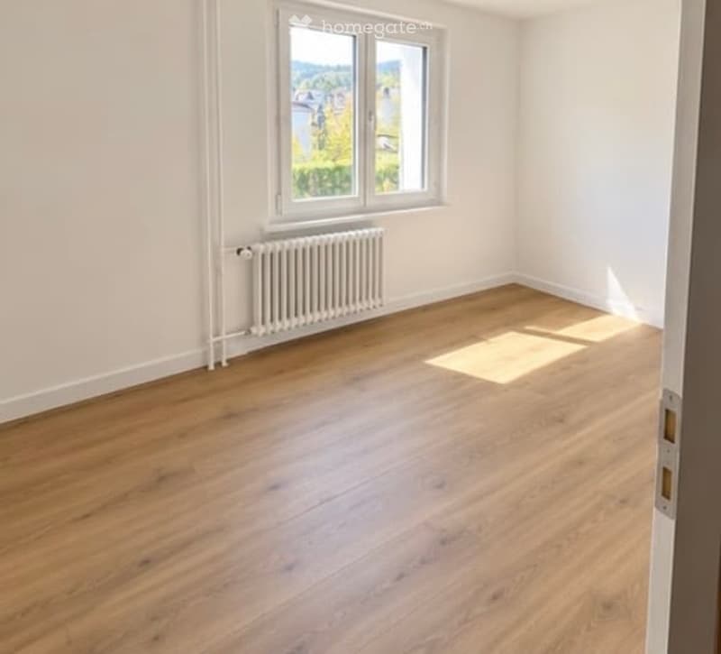 Empty room with wooden flooring, white walls, a radiator, and a large window with a view of greenery.