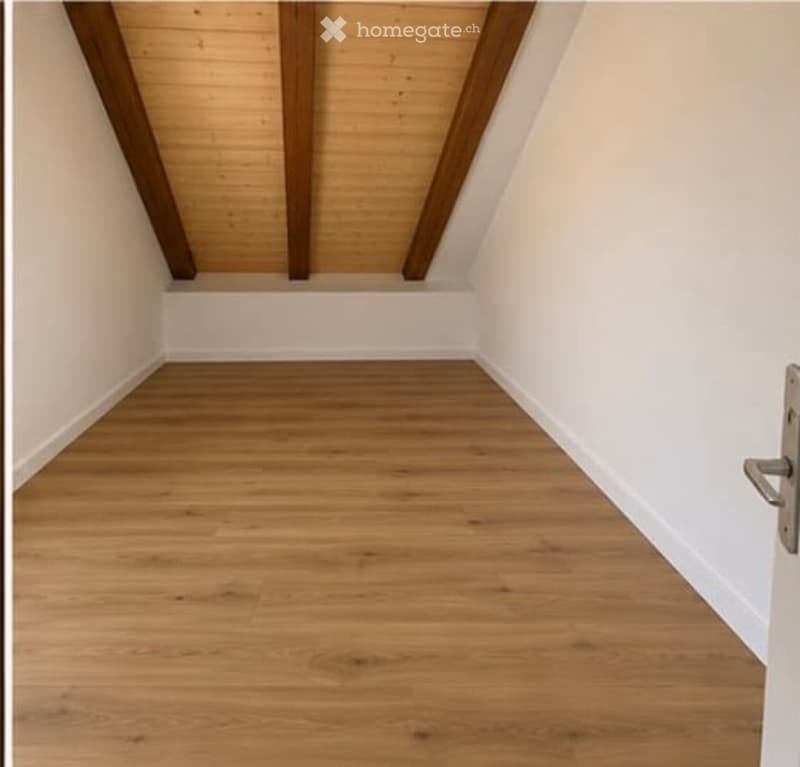 Empty room with wooden flooring and white walls, featuring exposed wooden beams on the ceiling and a door on the right side.