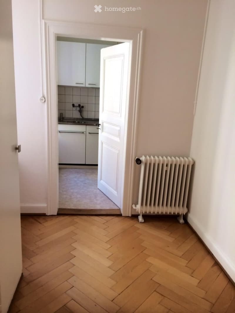 Room has parquet floor, white walls, an open door leading to a kitchen, a radiator