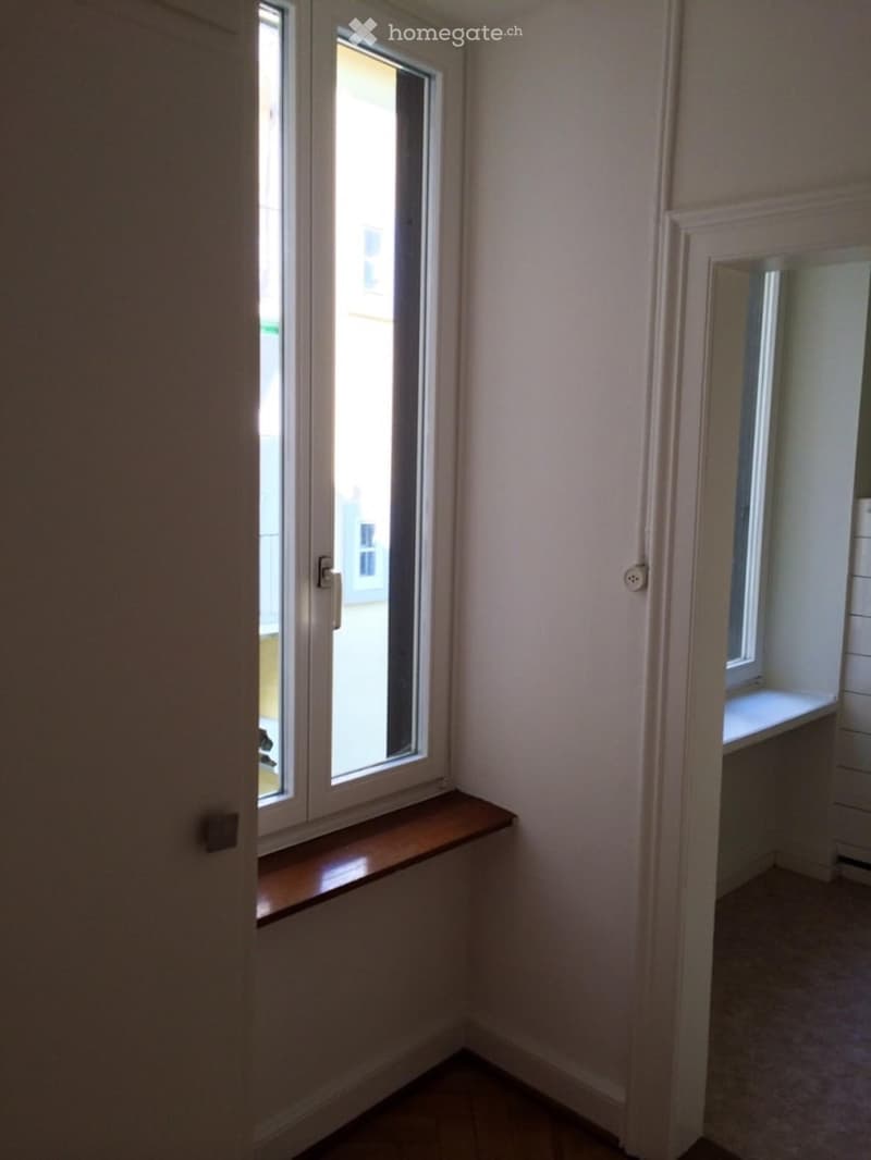 A narrow empty room with white walls, a wooden windowsill, and double windows with a view of a building outside.