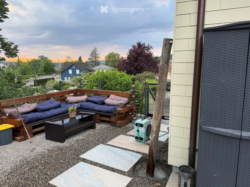 Outdoor patio with a wooden sofa, coffee table, small plant, and a fan. The area is surrounded by a fence and features a view of the neighborhood.