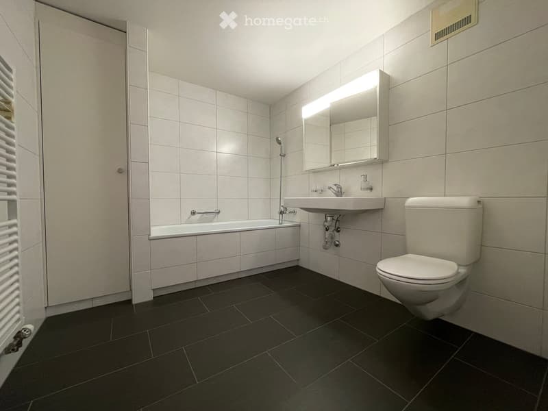 Modern bathroom with white tiles, dark floor tiles, bathtub, toilet, sink, and illuminated mirror.