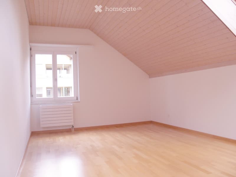 Empty room with light pink walls, wooden floor, and a window with white frame. The ceiling is slanted with wooden paneling.