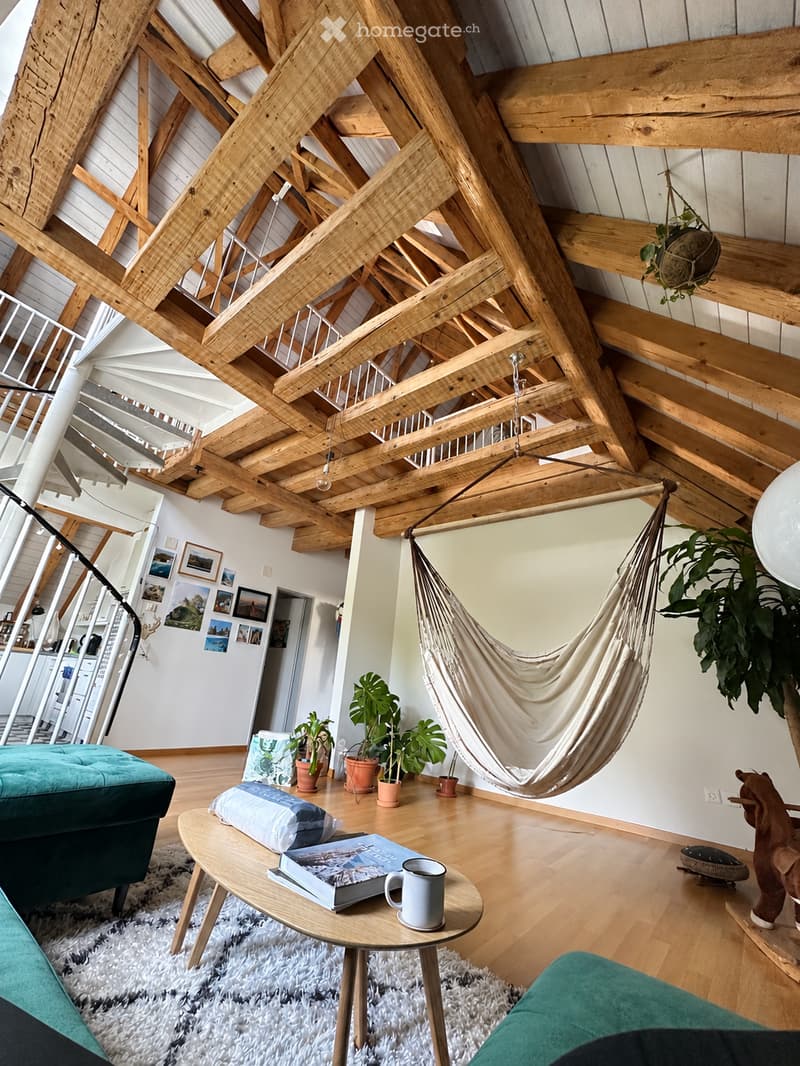 A modern living room with exposed wooden beams, a hanging hamaca, a teal sofa, a round coffee table with books and a mug, potted plants, and picture frames on the wall.
