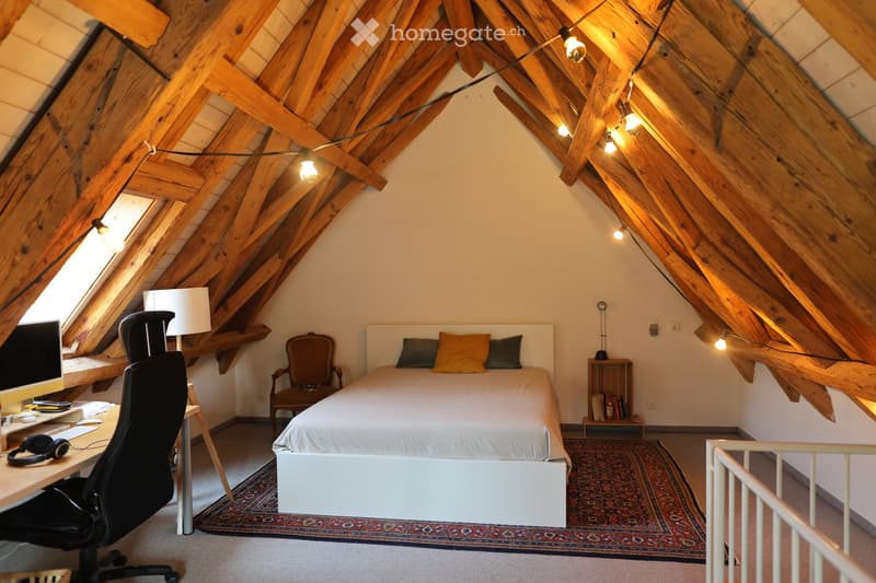 Cozy attic bedroom with wooden slanted ceilings, a white bed with pillows, a desk with a computer, a chair, and a small side table with books.