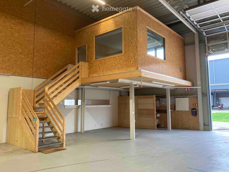 A modern, wooden structure with a raised ground floor and a staircase leading to an upper level, situated inside a larger industrial space with exposed metal beams and concrete floors.