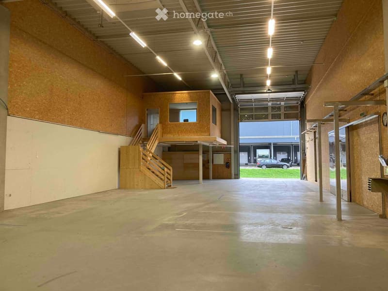 Empty industrial-style space with concrete floor, exposed wooden walls, metal ceiling, and a staircase leading to an upper level. Large garage door opens to an outdoor area with a parked car visible.
