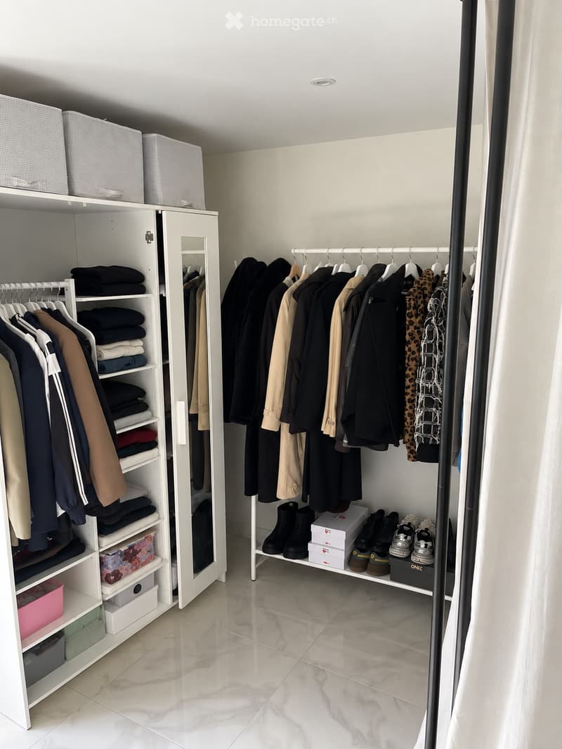 A well-organized walk-in closet with white built-in wardrobes, clothing neatly hung on racks, folded garments on shelves, and a shoe rack holding various footwear.