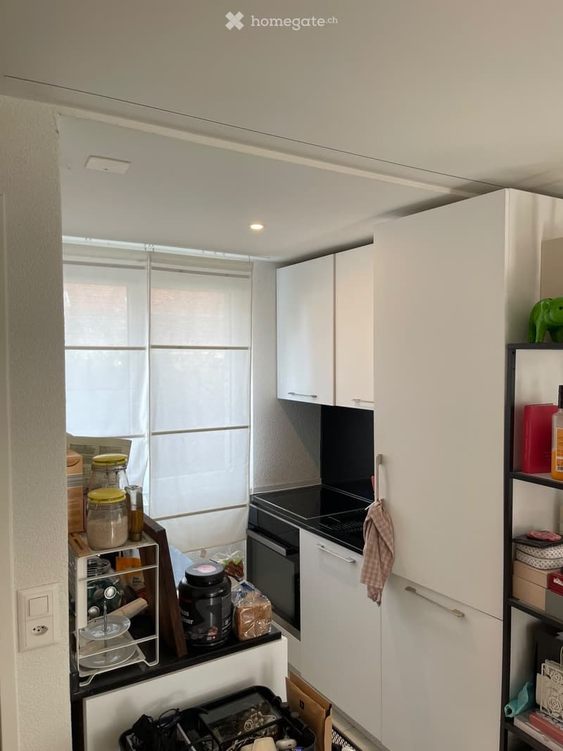Modern kitchen with white cabinets, stainless steel appliances, and a large window with blinds. Shelves hold jars, bottles, and other kitchen items.