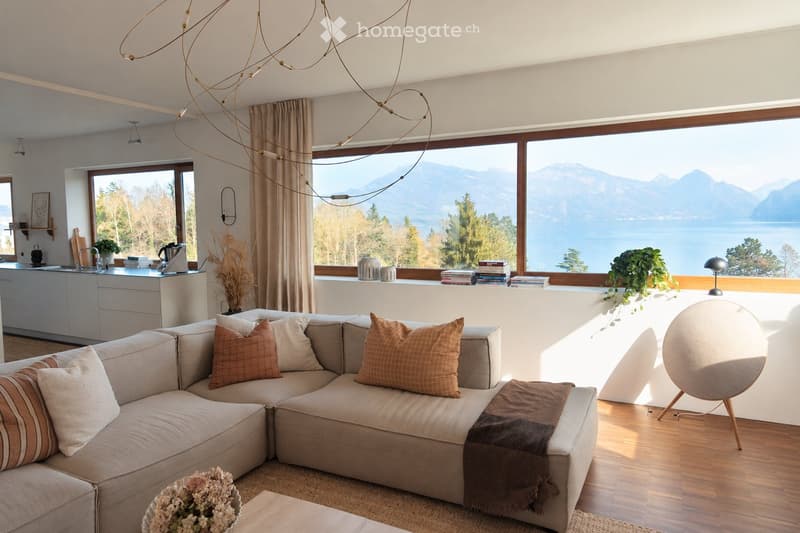 Modern living room with large windows offering a scenic view of a lake and mountains. Features a grey sectional sofa with throw pillows, a round wooden coffee table, a modern chair, and a chandelier with warm lighting. The kitchen area includes a counter,