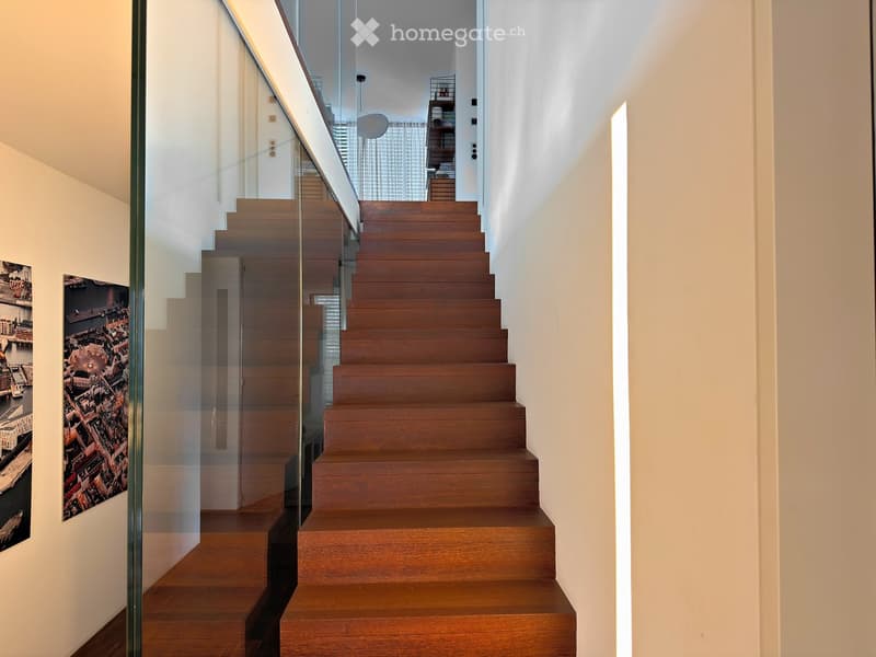 Modern wooden staircase with glass railing, white walls, and framed cityscape artwork on the left side.