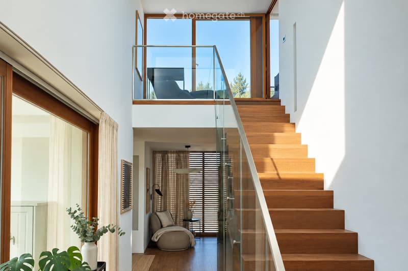 Modern hallway with wooden staircase, glass railing, large windows, and a cozy seating area visible in the background.