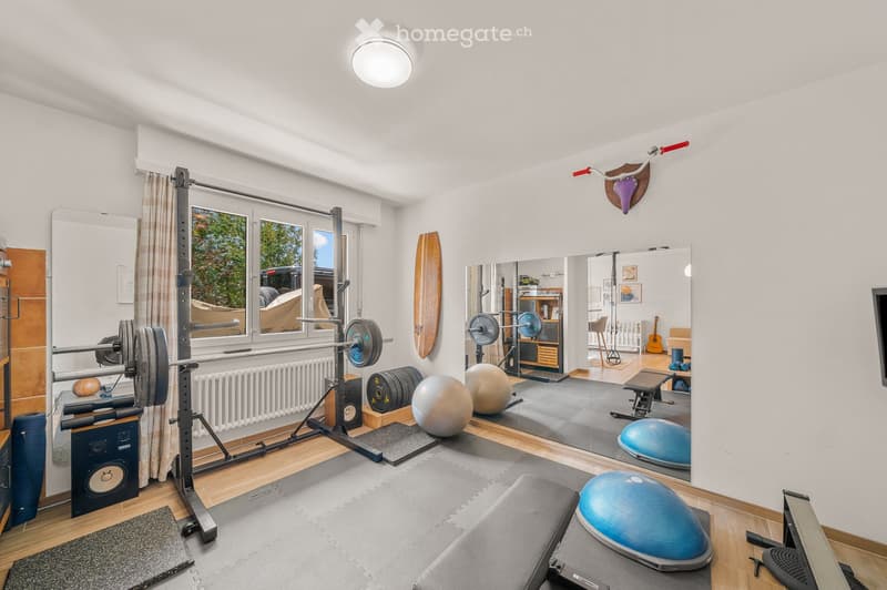 Well-equipped home gym with a barbell, weight plates, fitness balls, a large mirror, surfboard on the wall, and a guitar in the background.