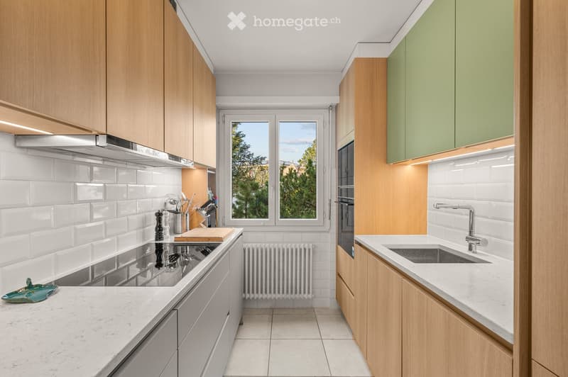 Modern kitchen with light wooden cabinets, white tiled walls, marble countertops, and stainless steel appliances. Features a sink, oven, and various kitchen utensils on the counter.