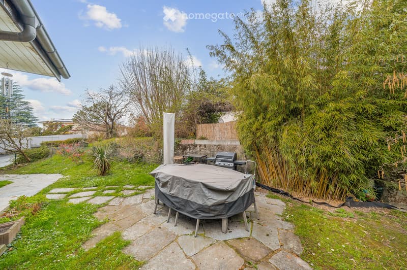 Garden area with a covered round table, barbecue, stone pathway, and surrounding greenery including bamboo plants and trees.