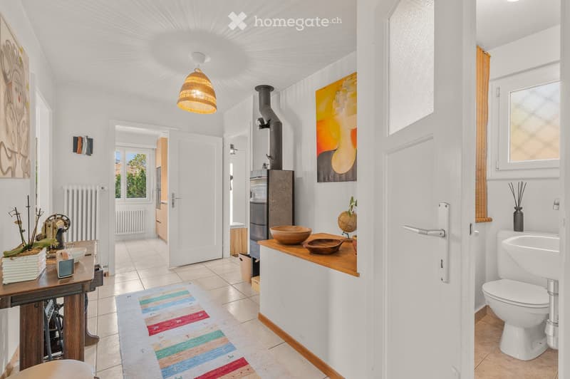 Bright hallway with white walls, tiled floor, and a hanging woven pendant light. Features a small wooden table with decorative items, a colorful runner, and an open doorway leading to another room with a visible radiator and artwork.