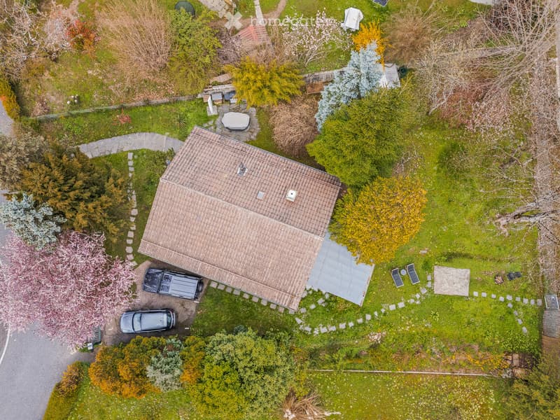 Aerial view of a two-story house with a tiled roof, surrounded by a well-maintained garden with trees, shrubs, and a paved driveway with parked cars.