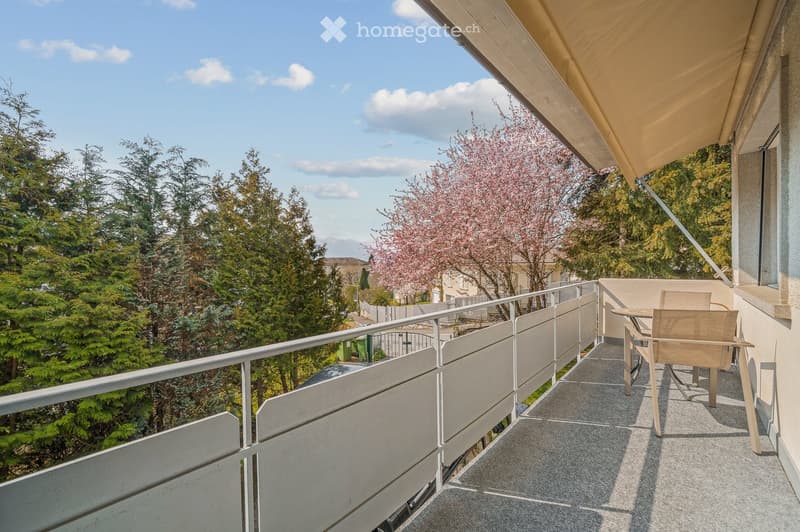 Balcony with a table and chair, overlooking a green area with trees and a flowering tree.