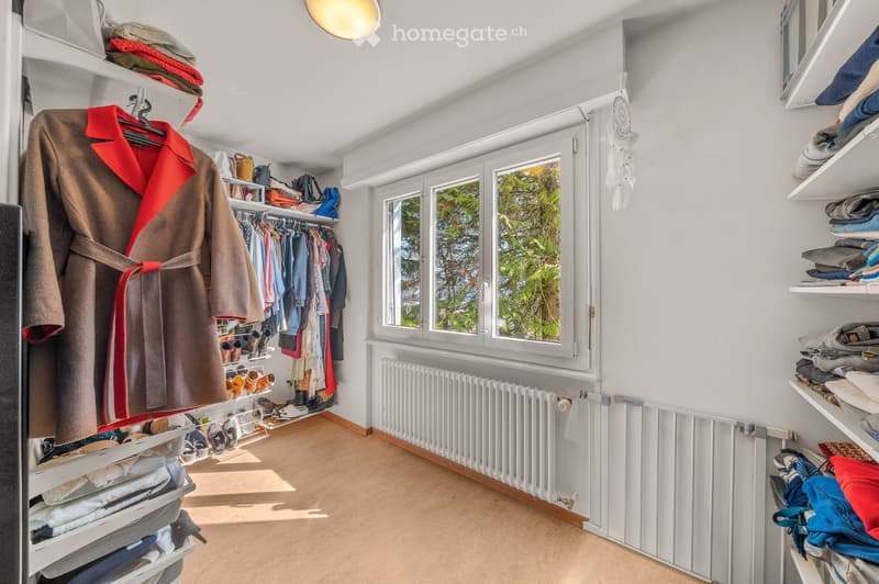 A well-organized walk-in closet with white walls, wooden flooring, and large windows. Features include a wardrobe with hanging clothes, shelves filled with folded garments, a coat on a hanger, a shoe rack, and a radiator below the windows.
