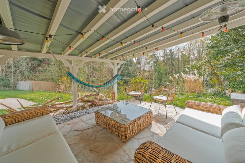 Outdoor patio with wicker furniture, glass coffee table, hammock, and string lights under a covered roof.