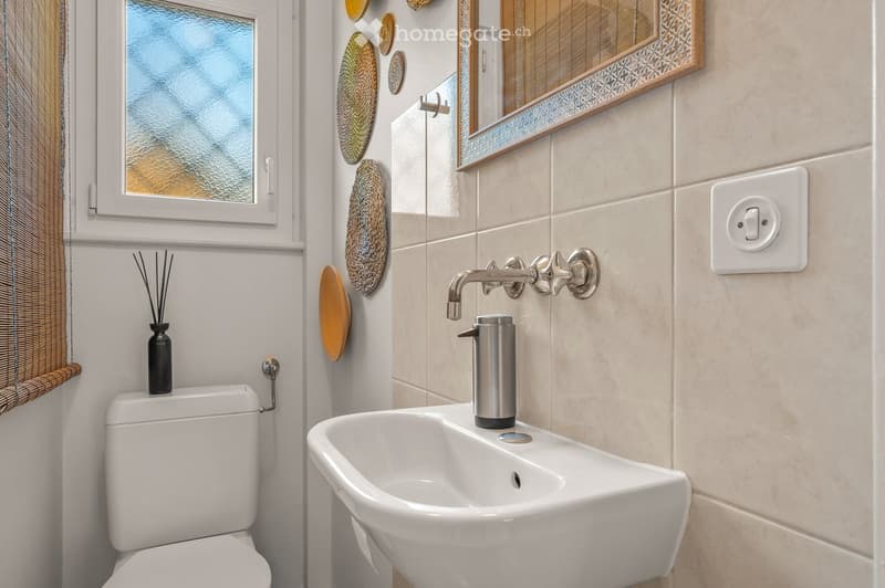 Modern bathroom with a white toilet, sink, and decorative plates on the wall. Features a large mirror with a wooden frame and a window with frosted glass.