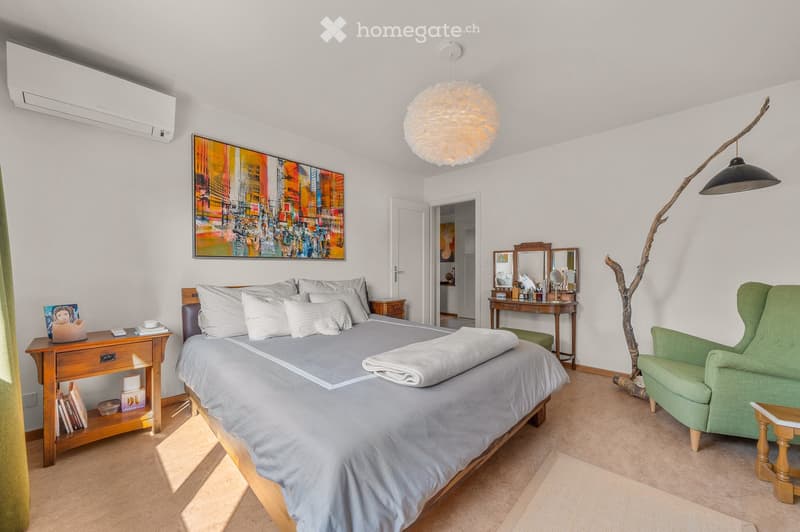 A well-furnished bedroom with a large bed, nightstand, green armchair, vanity table with mirrors, and a decorative floor lamp. The room features a colorful abstract painting on the wall and a small wooden shelf with books and decorative items.