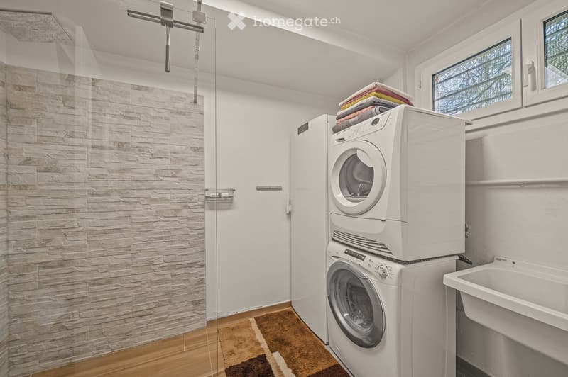 Modern bathroom with a glass-enclosed shower, white washing machine and dryer stacked, a sink, and a towel rack with folded towels.