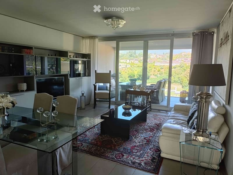 Elegant living room with large windows offering a scenic view, featuring a cream-colored sofa, glass coffee table, glass dining table with chairs, and a patterned area rug.