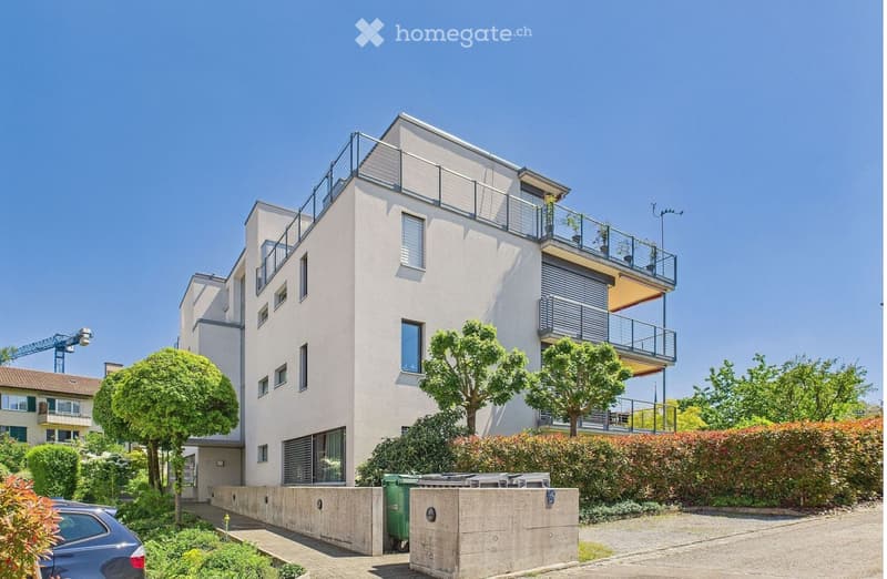 Modern multi-story building with balconies, large windows, and contemporary design, surrounded by greenery and a paved driveway.