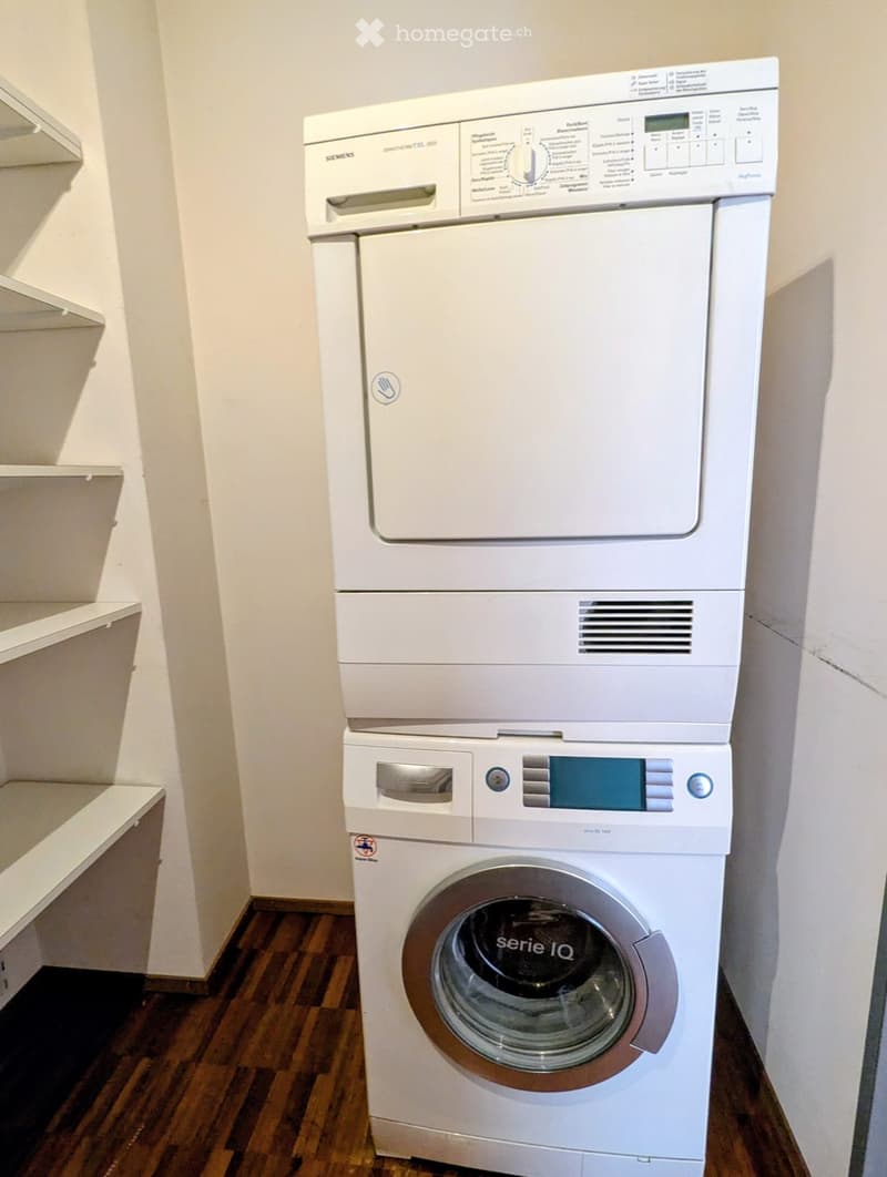 A compact bathroom with a stacked washing machine and dryer unit, featuring a wooden floor and white walls.