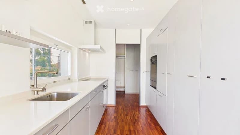 Bright, modern kitchen with white cabinets, a stainless steel sink, and a built-in oven. The space features a large window and a wooden floor.