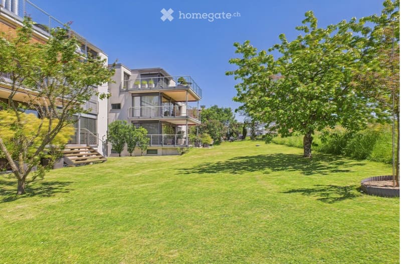Lush green lawn with trees and shrubs, modern house with balconies and stairs leading to the lawn.