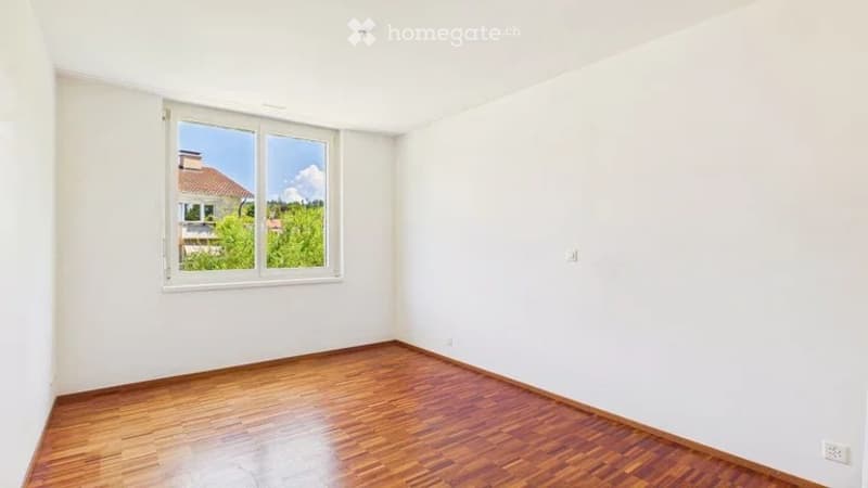 Empty room with wooden flooring and white walls, featuring a large window with white frame and view of outdoor area.