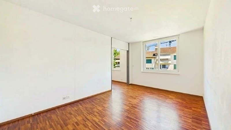 Empty room with wooden flooring and white walls, featuring a large window and a door leading to another space.