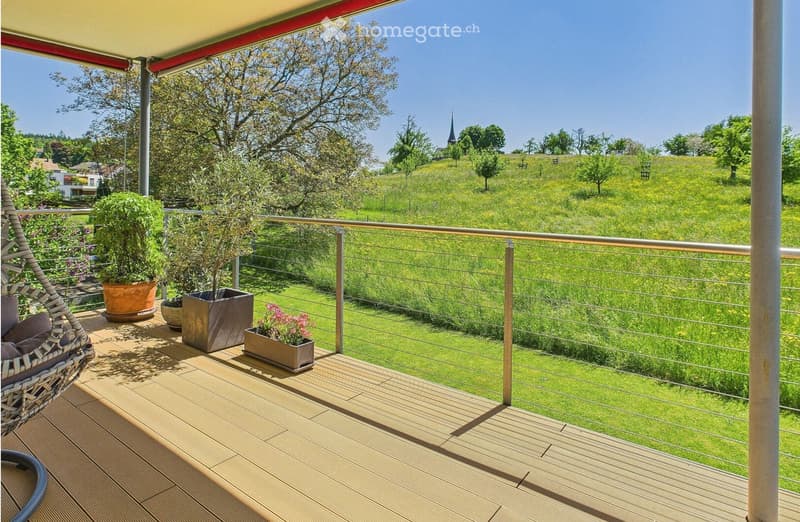 Balcony with wooden decking, metal railing, and potted plants, overlooking a green field and distant church spire.