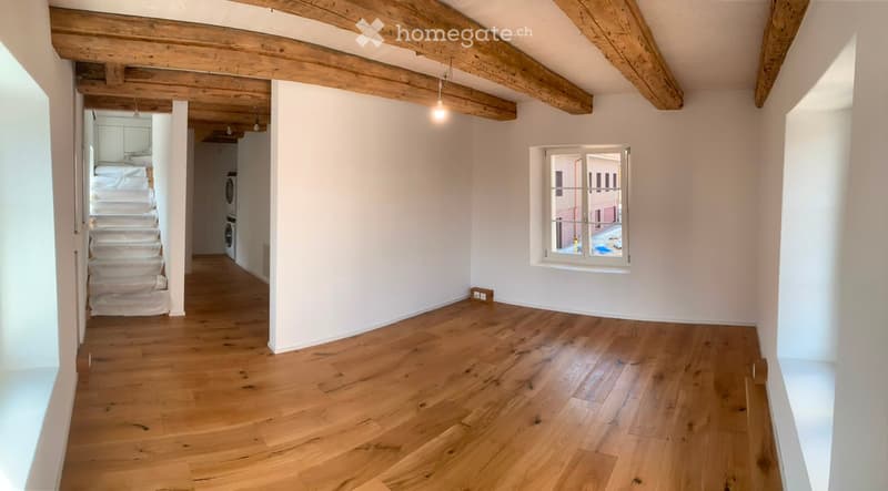 Empty room with wooden flooring, white walls, exposed wooden beams on the ceiling, and a window with a view of the street.