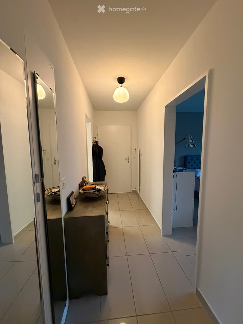 A narrow hallway with white walls, tiled floor, and a single hanging light fixture. There is a small console table with a mirror, a basket, and a framed picture on the left side, and an open doorway to a room on the right.