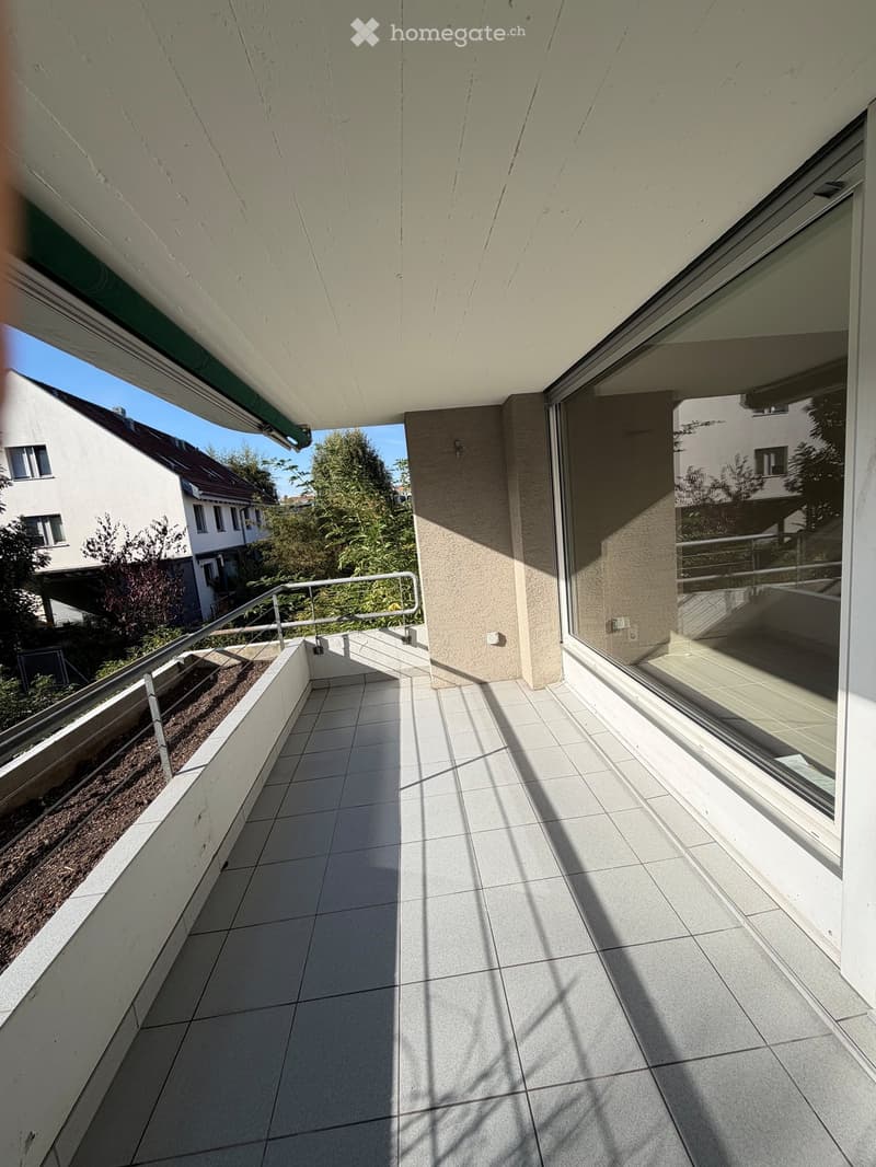 Balcony with tiled floor, metal railing, and large windows overlooking a residential area with trees and neighboring buildings.