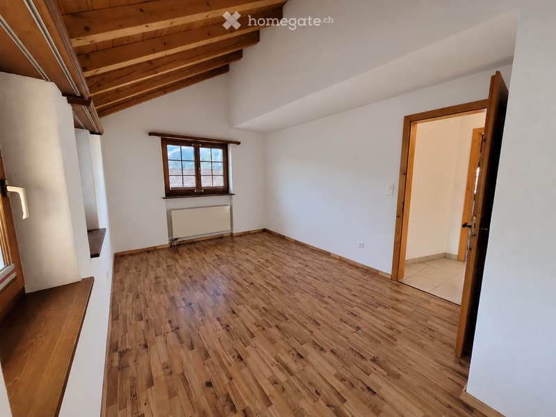 Empty room with wooden flooring, white walls, and a window with a view. The room features exposed wooden beams on the ceiling and a radiator below the window.