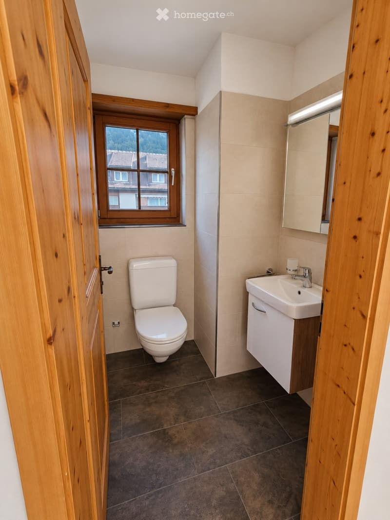 Modern bathroom with a toilet, sink, mirror, and cabinet. The room features tiled walls and a window with wooden framing.