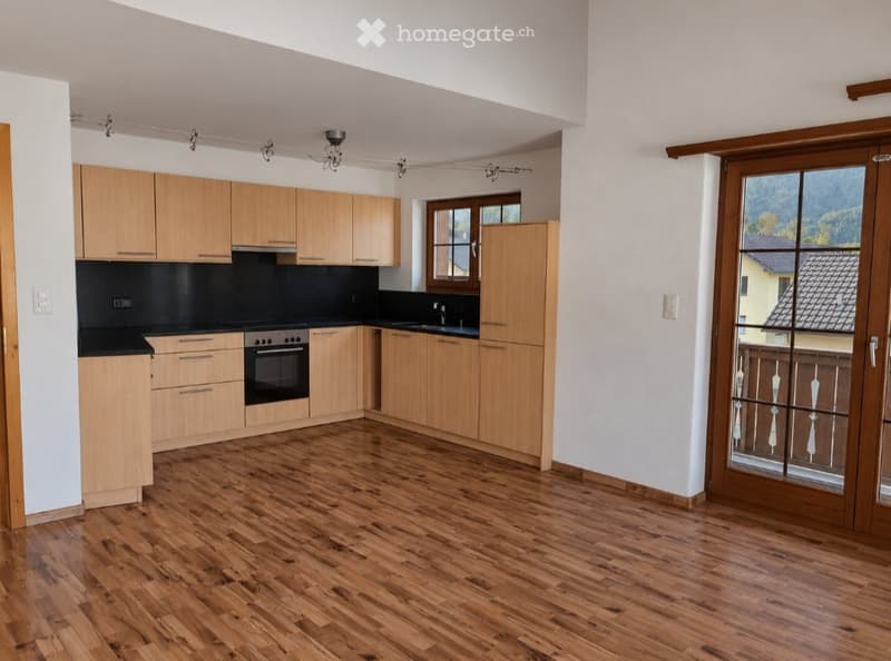 Modern kitchen with light wooden cabinets, black countertops, and a central island. Features include an oven, sink, and ample natural light from large windows and a sliding glass door leading to a balcony.