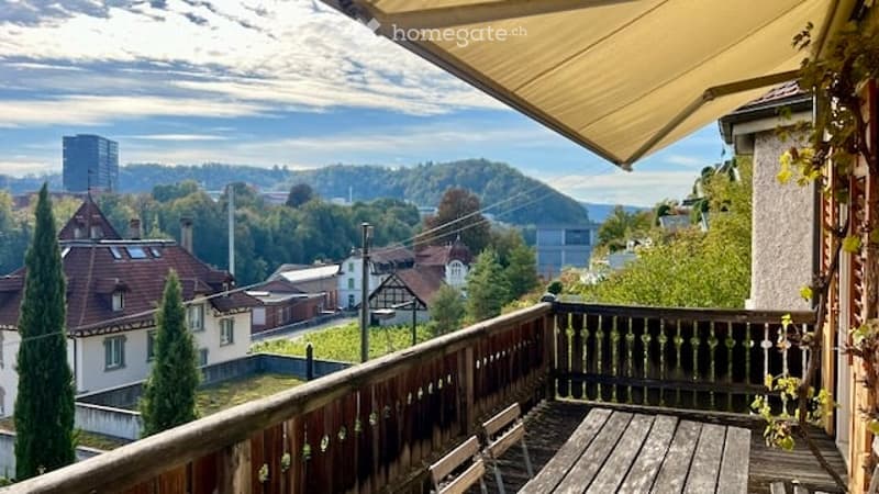 Balcony with wooden railing, table, and chairs, overlooking a scenic town view with houses and hills.