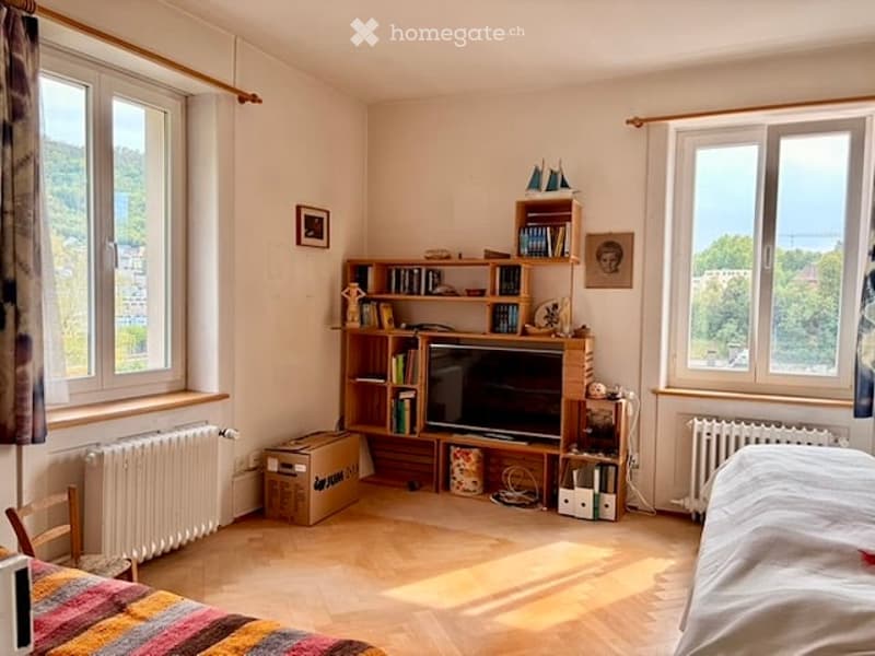 Cozy room with wooden flooring, large windows offering a scenic view, a bed with a colorful blanket, a bookshelf filled with books and decorative items, a television, and a radiator under the windows.