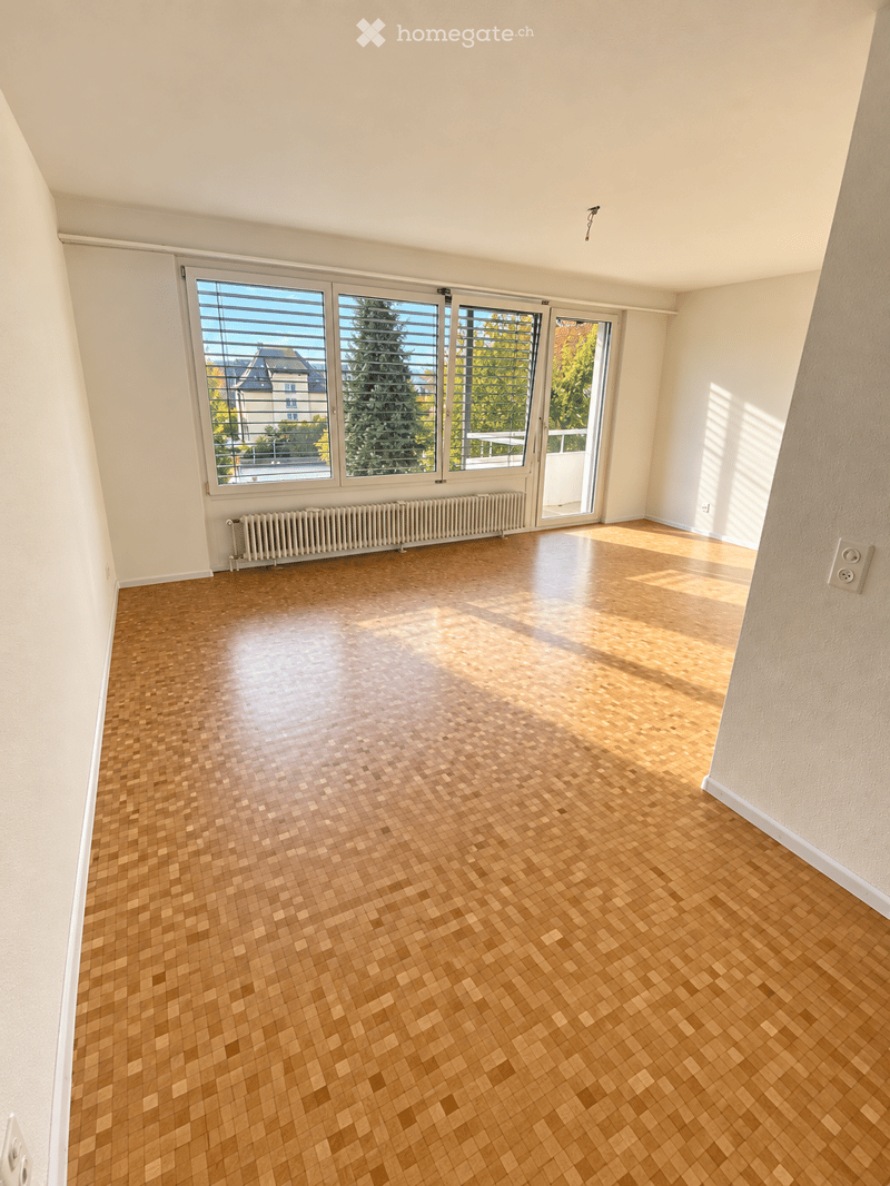 Empty room with wooden parquet flooring, white walls, large windows with blinds, radiator, and natural light streaming in.