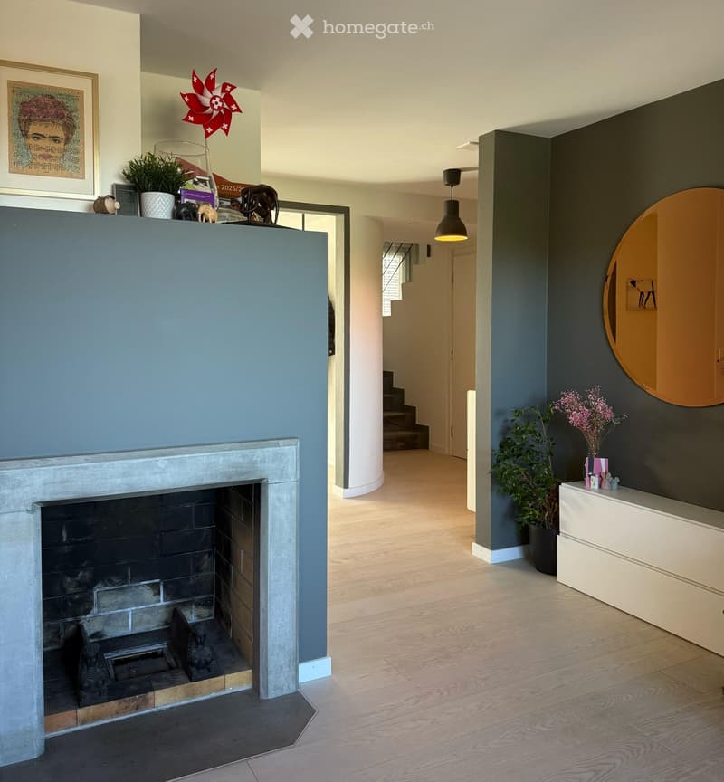 Modern hallway with a fireplace, wooden flooring, and a staircase leading upwards. Features include a round mirror, potted plants, and decorative items on a console table.