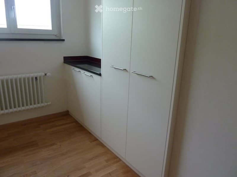 Empty room with white built-in wardrobes, wooden flooring, radiator, and a small window with a dark sill.